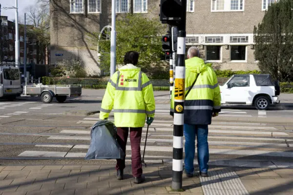 Twee werkgestraften in gele hesjes wachten bij een stoplicht. Een van de twee mannen heeft ene vuilniszak- en prikker bij zich.