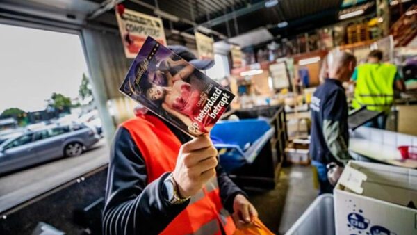 Een man staat in de werkplaats van een werkstraflocatie. Hij houdt een boek voor zijn gezicht om zijn identiteit te verbergen.