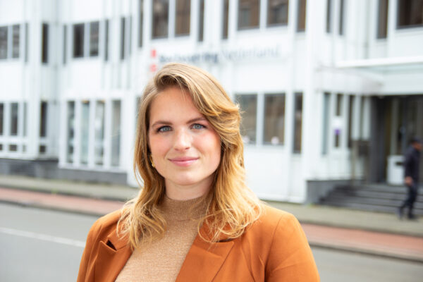 Portret van een jonge blonde vrouw met in de achtergrond een gebouw met het logo van Reclassering Nederland erop.